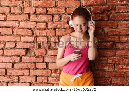 Photo of pleased caucasian woman with red hair using headphones and cellphone while standing isolated over brick wall indoors