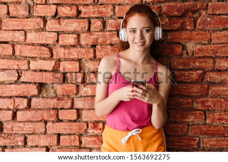 Photo of pleased young woman with red hair using headphones and cellphone while standing isolated over brick wall indoors