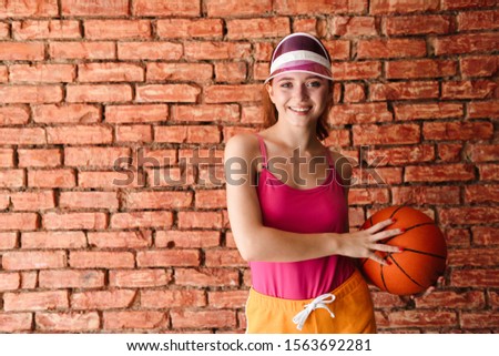 Photo of pleased young woman in visor holding basketball while working out isolated over brick wall indoors