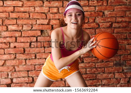 Photo of smiling young woman in visor playing basketball while working out isolated over brick wall indoors