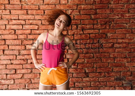 Image of a happy smiling amazing young redhead fitness woman posing over brick wall background shaking hair.