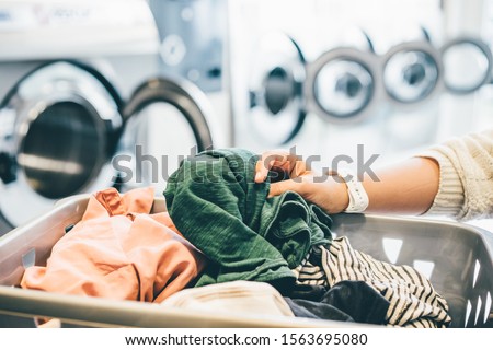 interior of small laundromat in daylight. Close-up female holding basket. Girl loading dirty clothes inside drum and closing door. Self-service concept Royalty-Free Stock Photo #1563695080