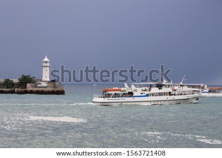 White tour vessel swimming in front of lighthouse in sea. Two boats carrying tourits for marine trip meet each other near shore on cloudy summer weather with pleaant sea breeze. Sea shore view image