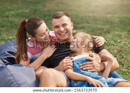 Picture of beautiful woman with daughter and husband sitting on blue frameless chair in park and enjoying to spend time together outdoors, wearing casual clothing. Happy family, relations concept.