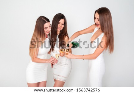 Three young woman in elegant dresses having fun, smiling, dancing and drinking champagne in studio on white background. Christmas party celebration concept.