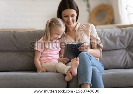 Smiling 30s woman holding digital tablet, sitting on cozy sofa with happy adorable little preschool daughter, watching funny cartoons or family photos, making purchases in online store, playing games.