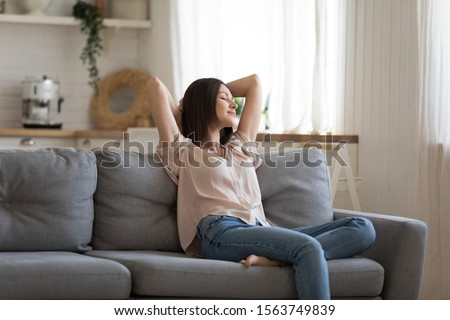 Happy young woman lying on comfortable couch in modern living room. Smiling lady crossing hand behind head, relaxing on cozy sofa at home after hard working day. 30s house wife accomplished chores. Royalty-Free Stock Photo #1563749839