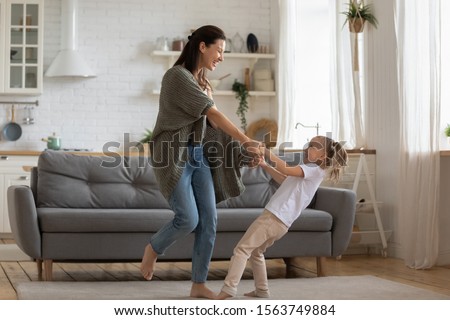 Overjoyed energetic small adorable daughter twisting with young mother. Happy 30s woman dancing to music with laughing little preschool daughter on carpet at studio kitchen, full body length image. Royalty-Free Stock Photo #1563749884