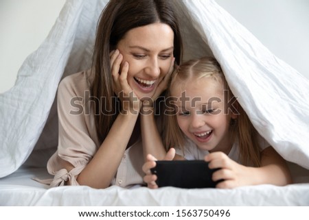 Head shot close up portrait happy young woman lying under blanket on bed with adorable overjoyed little daughter, playing online mobile games, watching funny videos, enjoying pastime together at home.