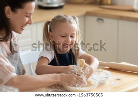 Head shot close up overjoyed little adorable daughter helping smiling young mother kneading dough by hands in bowl. Happy family in aprons enjoying pancakes bans pie cooking process at kitchen.