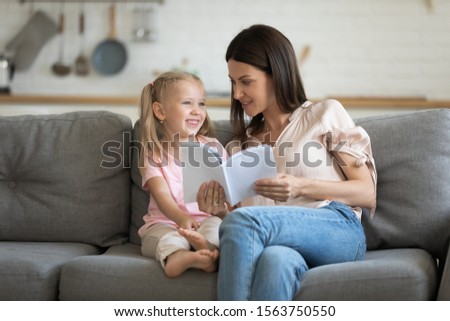 Happy little child girl sitting on sofa with attractive young mom, reading interesting books together. Smiling mother talking about story heroes with small adorable daughter, learning poems by heart.