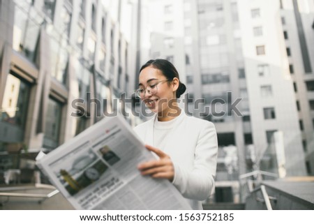 Young attractive Asian female banker or accountant in glasses is reading newspaper outside a modern office center or a bank Royalty-Free Stock Photo #1563752185