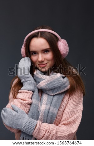 Portrait of young beautiful smiling girl in knitting pullover, mittens and pink fluffy earmuffs on a dark background. Xmas model with long straight hair. Winter holidays, Christmas, New Year concept.