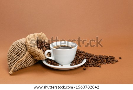 Hot espresso cup and coffee beans in burlap bag on brown background. White coffeecup or mug, whole roasted arabica coffea bean in natural jute rustic sack with copyspace Royalty-Free Stock Photo #1563782842