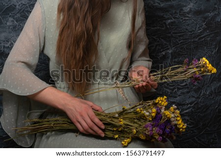 A girl in a beautiful vintage dress sits with a bouquet of immortals on her lap and counts them. Dramatic photo. Dark background
