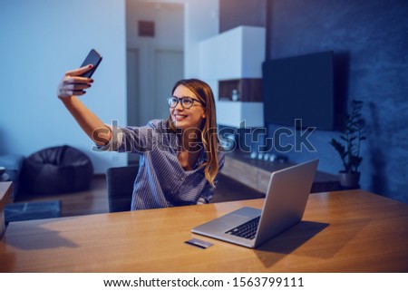 Young smiling charming Caucasian brunette in stripped shirt and with eyeglasses sitting at dining table and taking selfie. On table are laptop and credit card.