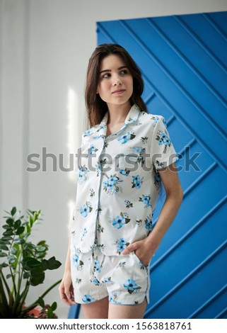 Young slim brunette woman, wearing white pajamas with light blue flowers pattern, posing for picture in front of blue wall and green plant in flowerpot. Sleepwear design. Cute girl in the morning