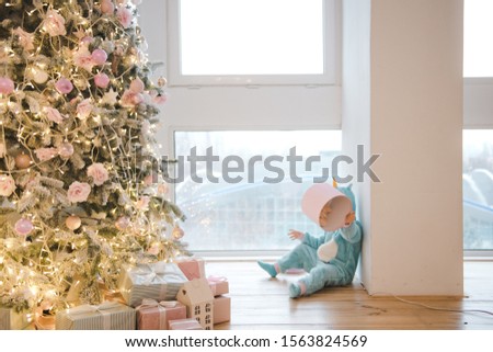 Child with a gift sits near a Christmas tree. Little baby clothing teddy bear sitting near the window. Christmas tree and gifts. New year home interior