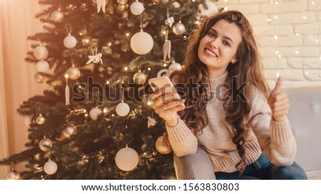 Woman with phone showing thumbs up sitting on the sofa near Christmas tree in the decorative interior. Positive gesture. Christmas and New Year photo. 