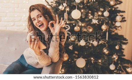 Woman with phone gesturing okay symbol sitting on the sofa near Christmas tree in the decorative interior. Positive gesture. Christmas and New Year photo. 