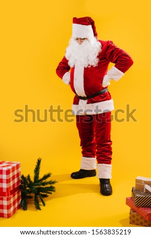 Puzzled look. Santa Claus putting hands on belt looks at gifts on the floor, isolated on yellow background