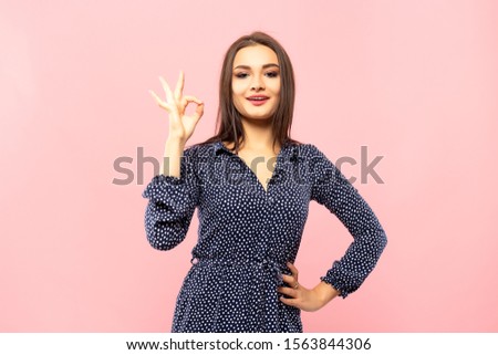 Cheerful pretty girl young cute beautiful woman in a blue dress with white polka dots shows ok gesture on pink background