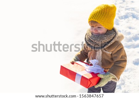 Kid in winter clothes holds Christmas gift. Kid during stroll in a snowy winter park. Beautiful winter nature. Amazing winter park. Adorable kid with cute face. Ptoto with empty copy space