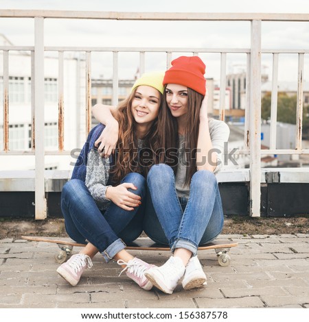 Two young girl friends sitting together on long-board and having fun. Outdoors, lifestyle.