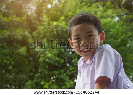 Smiled happily face of the child. Portrait of smiling Asian black hair boy age 6 years old with clean and healthy teeth wear a white shirt of kinder uniform on blurred green natural tree background.