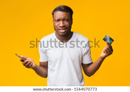 No Money. Confused Afro Guy Holding Smartphone And Credit Card Shrugging Shoulders Posing In Studio Over Yellow Background.
