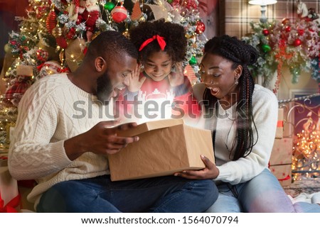 Magical Christmas. Excited african american family of three opening shining gift box in living room near Xmas tree, celebrating holidays at home
