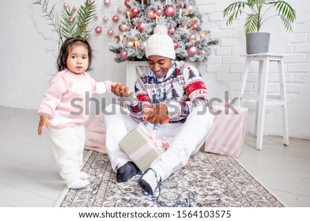 Young african-american father hugs his little charming daughter sitting in living room on the background of Christmas tree LED lights and gifts. Concept of a family holiday and gifts for children