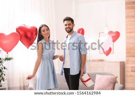 Young man presenting gift to his girlfriend in living room decorated with heart shaped balloons. Valentine's day celebration