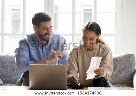 Head shot happy young family couple planning monthly budget, sitting together on cozy sofa, using computer software. Smiling mixed race wife calculating income and outcome together with husband. Royalty-Free Stock Photo #1564174900
