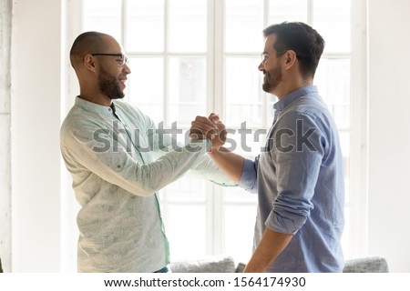 Side view excited mixed race male friends in eyewear shaking hands. Happy best buddies welcoming each other at sudden meeting. Smiling coworkers saying hi, communicating together indoors. Royalty-Free Stock Photo #1564174930