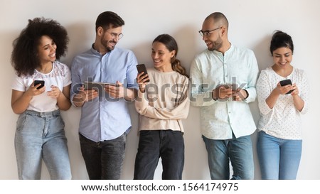 Happy diverse young people standing in row isolated on white background, using different electronic devices. Obsessed with technology smiling mixed race students friends sharing information. Royalty-Free Stock Photo #1564174975