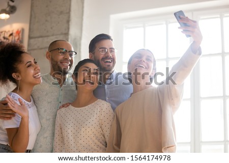 Head shot happy mixed race girl taking selfie photo of smiling young multiracial friends, looking at camera. Group of laughing students having fun together, recording funny video on smartphone.