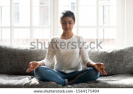 Full length mindful young indian woman making mudra gesture, sitting in lotus position on comfortable couch at home. Peaceful millennial girl deeply meditating, doing breathing yoga exercises alone. Royalty-Free Stock Photo #1564175404