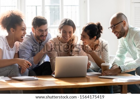 Smiling young mixed race woman showing funny videos on computer to happy multiracial friends, sitting together on cozy couch at home. Overjoyed diverse millennial people having fun in dormitory. Royalty-Free Stock Photo #1564175419