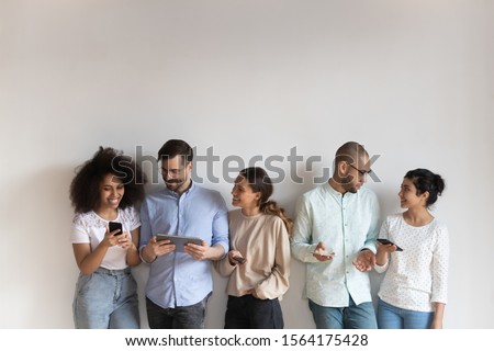 Group of smiling young mixed race people standing near white wall in row with different gadgets. Happy multiracial friends communicating, talking, sharing contacts or information, discussing apps.