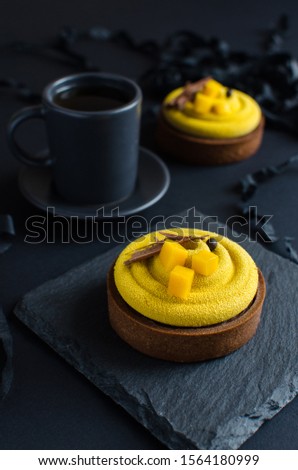 Contemporary chocolate mango mini mousse tarts covered with yellow velvet spray decorated with peace of fresh mango and cup of black tea on black background