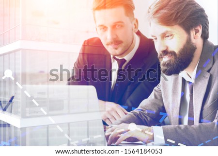 Two happy young businessmen working with laptop in abstract city with double exposure of graph. Concept of teamwork and corporate lifestyle. Toned image