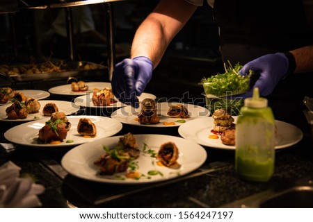 Chef's hands in nitrile gloves plates up food on the pass. Gloves are lilac in colour. White plates with colourful food arranged in a decorative fashion. Dark background with warm toned light. Royalty-Free Stock Photo #1564247197