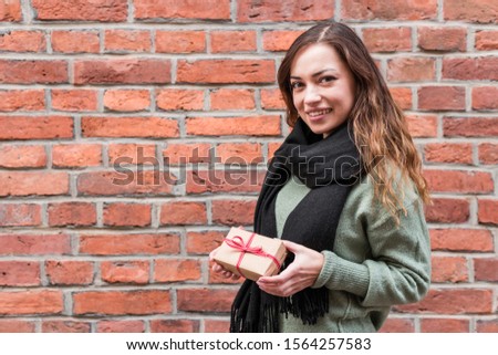 Cheerful young woman holding small gift box for Christmas or Valentine Day wrapped in brown paper and red ribbon. Copy space. Brick old background.