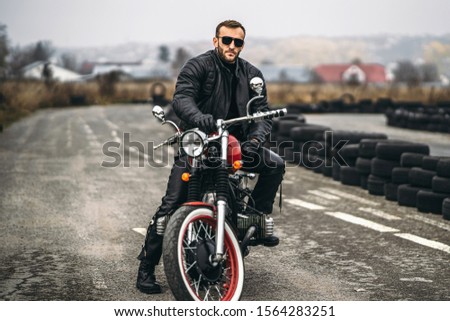 Bearded man in sunglasses and leather jacket looking at the camera while sitting on a motorcycle on the road. Behind him is a row of tires.