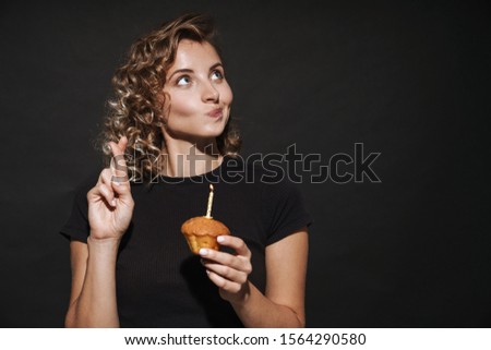 Image of pretty hopeful young woman isolated over dark grey wall background holding birthday cupcake with candle make fingers crossed gesture.