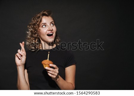 Image of surprised happy hopeful young woman isolated over dark grey wall background holding birthday cupcake with candle make fingers crossed gesture.