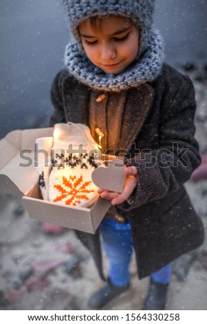 Little pretty girl in knitted grey hat opening a crafted gift box with a new pair of gloves. Baby, it is cold outside, white snowfall and cold weather, outside lifestyle portrait, focus on gloves