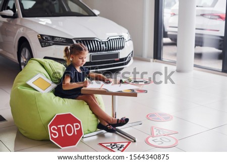 Cute little girl sits on the soft green chair by the table with pencil and paper sheets. Near modern automobile and road signs on the floor.
