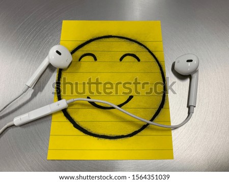 Drawing smile face on yellow sticky paper note and white earphone on silver background.
